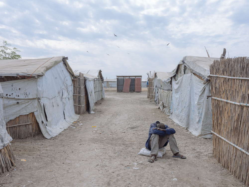 Bentiu, South Sudan, 2018. A man sits between shelters in Bentiu PoC (Protection of Civilians) site. [ Emin Ozmen/Magnum Photos ]
