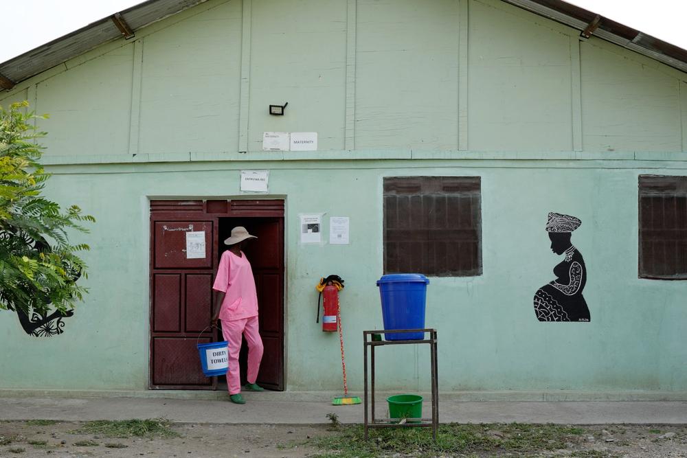 The maternity ward at MSF’s Ulang Hospital in South Sudan, featuring a mural of a pregnant woman on a pale green wall. A staff member in pink scrubs exits, carrying a bucket labeled “Dirty Towels.